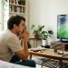 Homem de camiseta bege sentado em sofá cinza com mãos no queixo olhando tenso para TV com partida de futebol.
