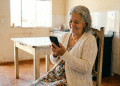 Idosa sorridente usa celular em casa simples.