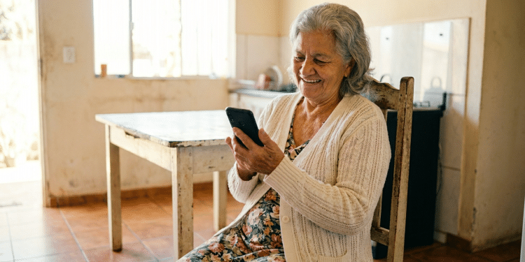 Idosa sorridente usa celular em casa simples.