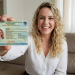 Mulher loira com cabelos cacheados mostra Carteira de Identidade Nacional do Brasil sentada em sofá.