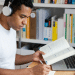 Homem jovem com fone de ouvido estudando com livro aberto e notebook em mesa de madeira.