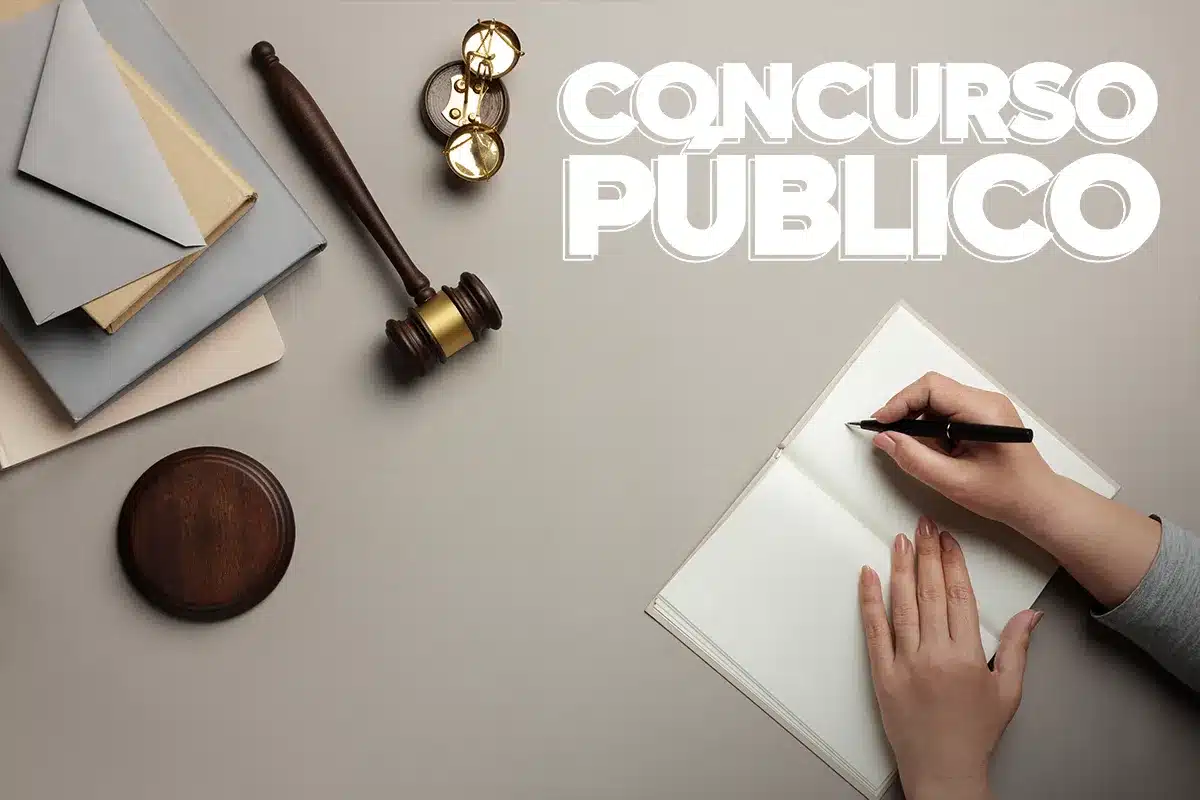Mãos escrevendo em caderno ao lado de martelo de juiz, livros e envelope, texto "CONCURSO PÚBLICO".Mãos femininas escrevem em caderno aberto sobre mesa cinza, próximos a livros, envelope, martelo de juiz de madeira e óculos, com texto "CONCURSO PÚBLICO" destacado no canto superior direito, simbolizando estudo e preparação para concursos jurídicos.