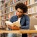 Jovem lendo livro em biblioteca rodeado por tecnologia e livros.