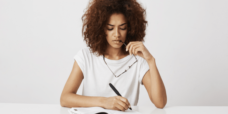 Jovem mulher refletindo enquanto escreve em caderno branco minimalista.