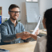 Homem de óculos sorri enquanto aperta a mão de uma mulher e segura currículos em ambiente corporativo.