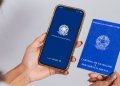 Mulher segura celular com aplicativo Carteira de Trabalho Digital aberto e carteira física azul ao lado.