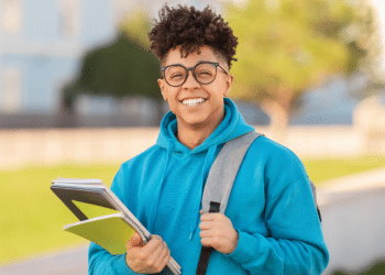 Jovem sorrindo com cadernos em campus universitário ao ar livre.