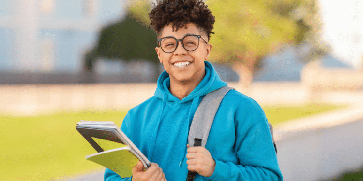 Jovem sorrindo com cadernos em campus universitário ao ar livre.