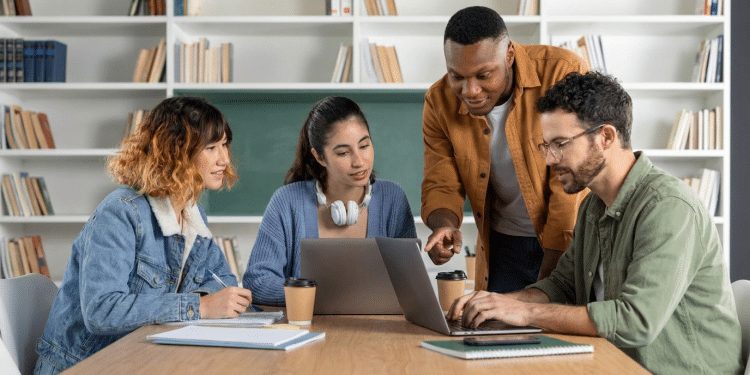 Quatro estudantes reunidos em mesa de biblioteca, um aponta para laptop enquanto outros observam com cadernos e café.