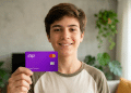 Jovem sorridente exibe cartão Nubank em casa iluminada.