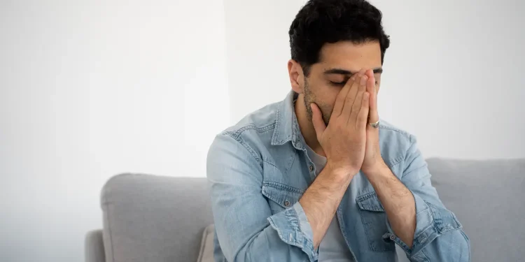 Homem em ambiente doméstico demonstrando sinais de ansiedade e tensão emocional ao cobrir o rosto com as mãos.