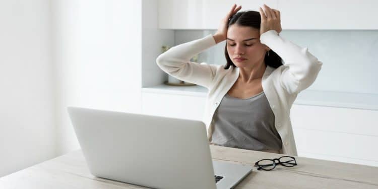 Mulher com as mãos na cabeça diante do notebook representa o estresse e o esgotamento causados pela autocobrança