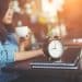 Mulher toma café ao lado de relógio despertador e notebook sobre mesa de trabalho