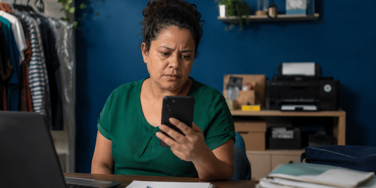 Microempreendedora consultando benefícios do INSS pelo celular em pequeno ambiente de trabalho.