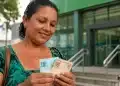 Mulher segura cédulas de real em frente a uma unidade da Caixa, representando o pagamento do Bolsa Família de junho.