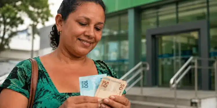 Mulher segura cédulas de real em frente a uma unidade da Caixa, representando o pagamento do Bolsa Família de junho.