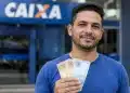 Homem brasileiro em frente à agência da Caixa Econômica Federal segurando dinheiro, representando liberação de saque de até R$ 6.220.