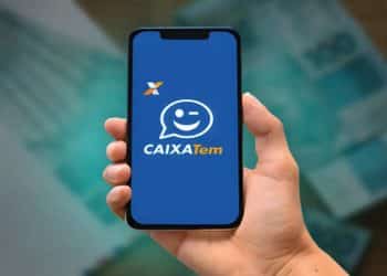 Celular exibindo a tela inicial do aplicativo Caixa Tem, segurado por uma mão, com notas de real desfocadas ao fundo, representando o acompanhamento de benefícios e pagamentos.