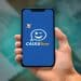 Celular exibindo a tela inicial do aplicativo Caixa Tem, segurado por uma mão, com notas de real desfocadas ao fundo, representando o acompanhamento de benefícios e pagamentos.