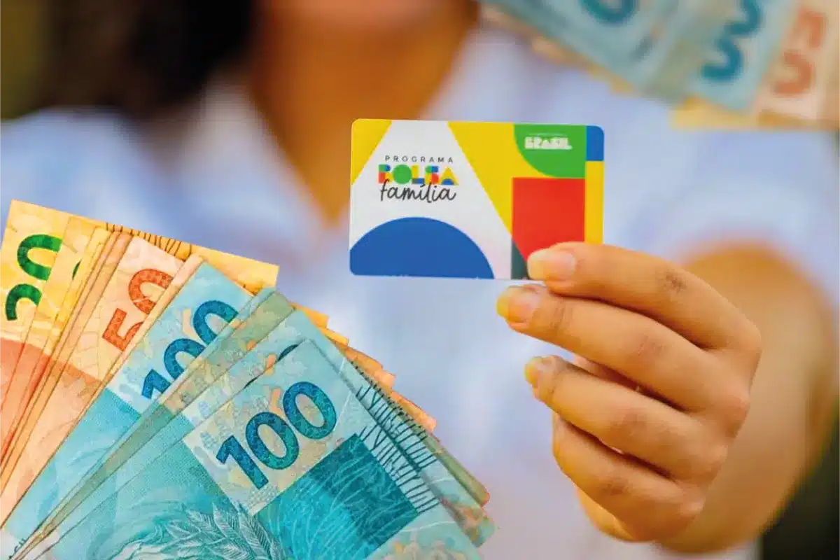 Mão segurando o cartão do Programa Bolsa Família ao lado de cédulas de R$ 50 e R$ 100, em referência ao calendário de pagamento de junho de 2026