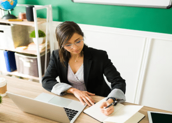 Mulher de óculos e blazer escreve em caderno ao lado do notebook