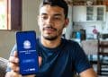 Homem sentado na cozinha de casa mostra para a câmera o celular com a tela inicial do aplicativo oficial da Carteira de Trabalho Digital, exibindo o botão Entrar e o brasão do Ministério do Trabalho