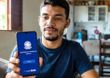 Homem sentado na cozinha de casa mostra para a câmera o celular com a tela inicial do aplicativo oficial da Carteira de Trabalho Digital, exibindo o botão Entrar e o brasão do Ministério do Trabalho