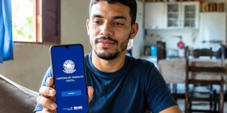 Homem sentado na cozinha de casa mostra para a câmera o celular com a tela inicial do aplicativo oficial da Carteira de Trabalho Digital, exibindo o botão Entrar e o brasão do Ministério do Trabalho