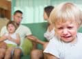 Criança pequena chora em primeiro plano enquanto a família aparece desfocada ao fundo