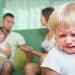 Criança pequena chora em primeiro plano enquanto a família aparece desfocada ao fundo