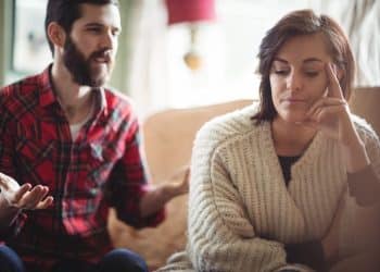 Mulher pensativa e cabisbaixa ao lado de homem que fala gesticulando no sofá, representando jogos mentais em uma conversa