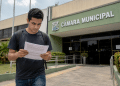 Pessoa lendo documento de concurso público em frente a prédio da Câmara Municipal.