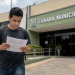 Pessoa lendo documento de concurso público em frente a prédio da Câmara Municipal.