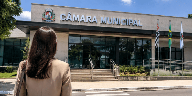 Pessoa em frente à Câmara Municipal representando divulgação de concurso público com vagas para níveis fundamental e médio.