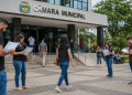 Pessoas em frente à Câmara Municipal lendo documentos de concurso público durante o período de inscrições abertas.