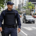 Guarda municipal de uniforme azul patrulha calçada de avenida movimentada, prédios ao fundo.