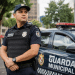 Guarda Municipal de uniforme azul-marinho com colete e rádio comunicador ao lado de viatura oficial.