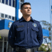 Guarda civil municipal de uniforme azul marinho com brasão, cinto com fivela, em frente a prédio azul e bandeira do Brasil.