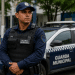 Guarda Municipal com uniforme azul marinho, colete tático e rádio comunicador posicionado ao lado de viatura.