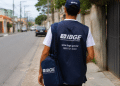 Agente do IBGE caminha por rua residencial com colete e bolsa institucional, representando atuação em campo no concurso para Agente Operacional.
