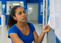 Mulher observa atentamente lista impressa afixada em mural azul em ambiente institucional.