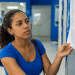 Mulher observa atentamente lista impressa afixada em mural azul em ambiente institucional.