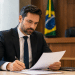 Homem de terno azul marinho e gravata assina documento em mesa de escritório, com bandeira do Brasil ao fundo.