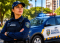 Guarda Municipal de braços cruzados, uniforme azul marinho com colete e boné, ao lado de viatura branca e azul com giroflex.