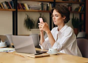 Mulher trabalha em home office com notebook sobre a mesa enquanto utiliza o celular como apoio à conexão de internet