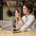 Mulher trabalha em home office com notebook sobre a mesa enquanto utiliza o celular como apoio à conexão de internet.