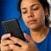 Mulher de camiseta azul consulta atenta a tela do celular para verificar a restrição de um aparelho pelo IMEI.