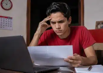 Jovem estudante brasileiro concentrado em notebook, em ambiente doméstico simples, com expressão de atenção e preocupação, representando a inscrição no Enem 2026 antes do prazo final.