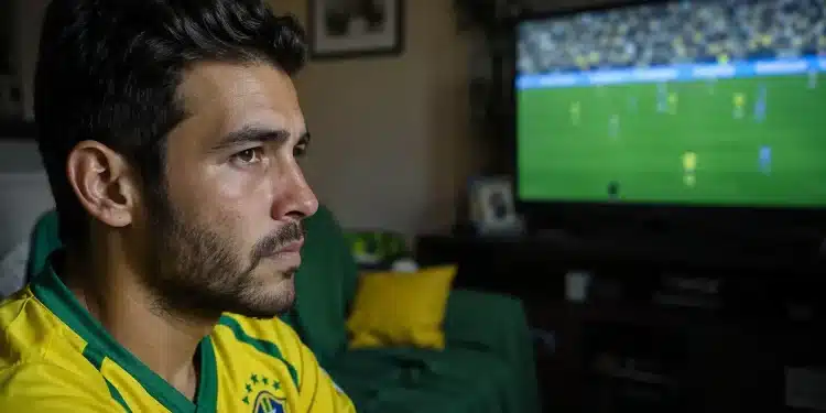 Jovem brasileiro assiste a jogo da Copa do Mundo na TV com expressão de cansaço visual.