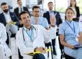 Profissionais de saúde de jaleco assistem a uma palestra, um deles com o braço levantado para participar